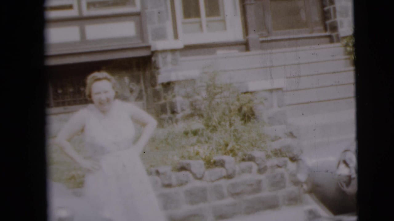 Woman in a light dress standing by a low stone wall near a parked car.