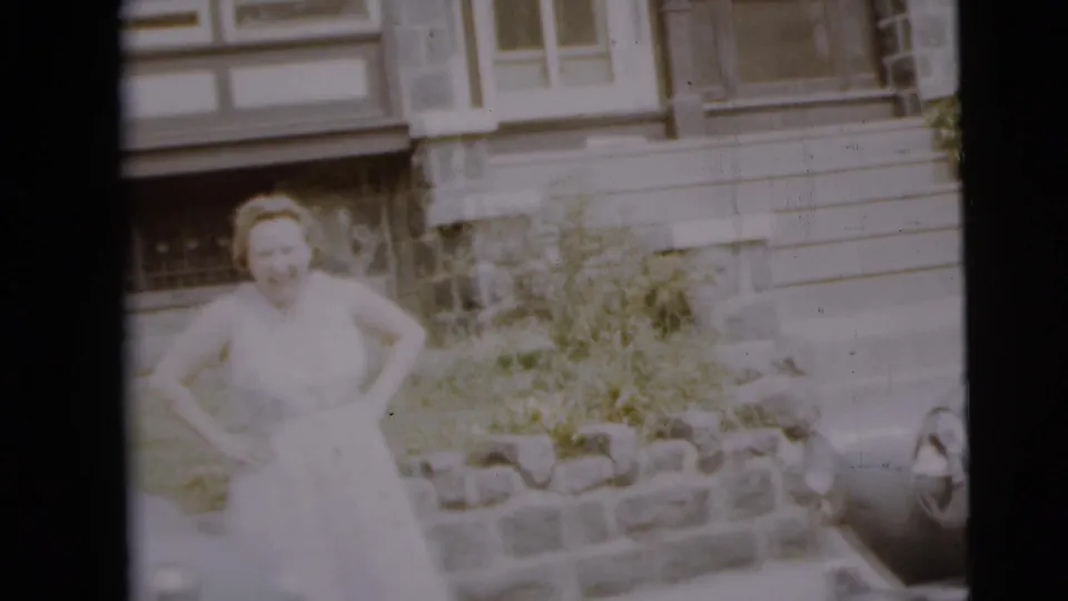 A woman in a light dress poses near a low stone wall outside a Camden residence in the 1962 home movie.