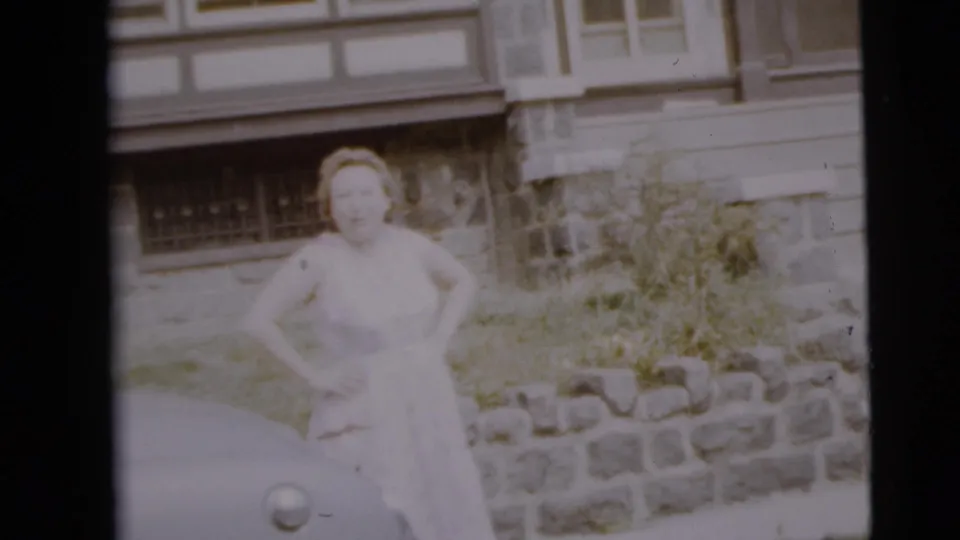 The woman remains posed outside the Camden residence, with an older car edge and garden stonework visible.
