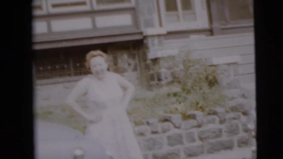 A soft 8mm frame shows the woman standing by shrubs and a stone wall outside the Camden residential building.