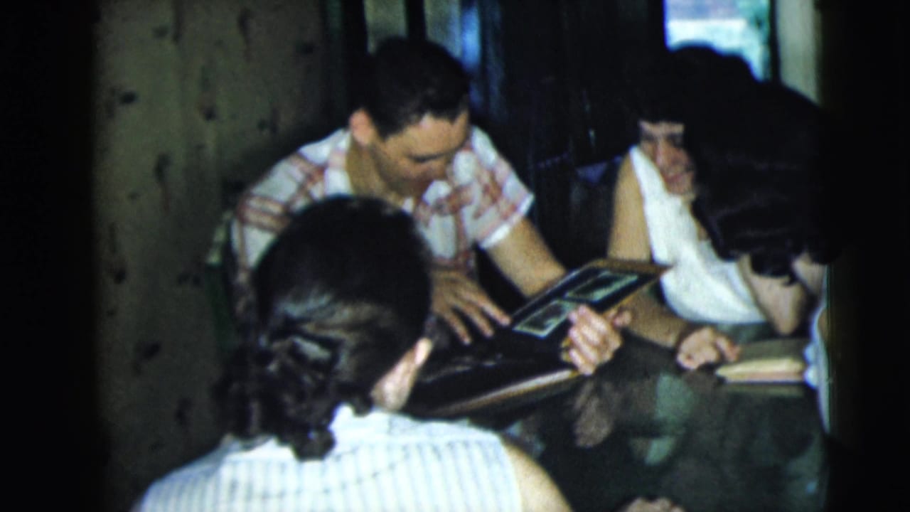 A man shows album pages to women seated around a table.