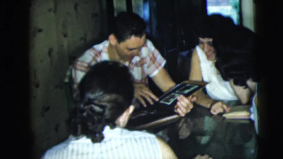 Adults in a dim Camden interior look toward a small photo album as the man in a checkered shirt leans forward.