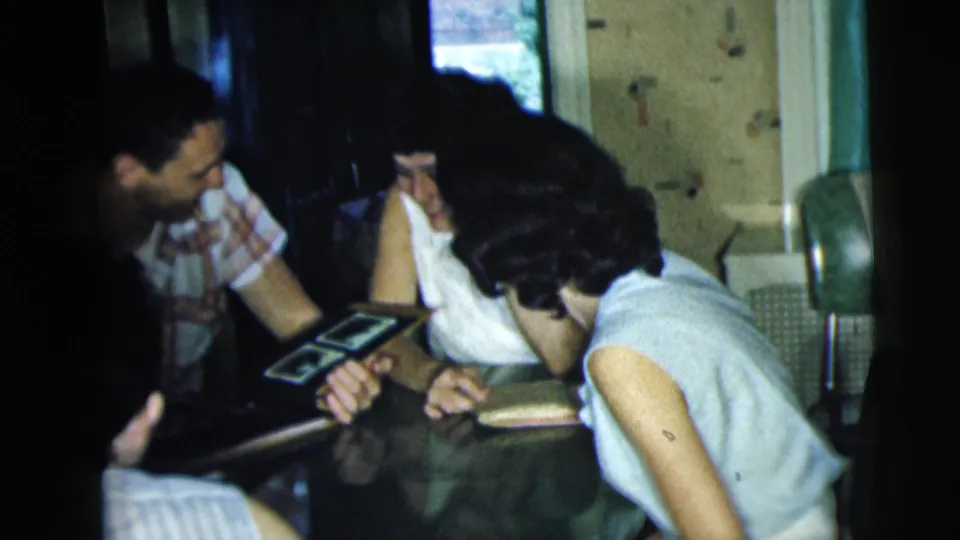 The group bends closer over the table, with the album open and a woman in a sleeveless top leaning in to see.