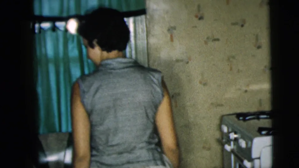 In a 1962 Camden home interior, a woman in a sleeveless top enters a room near green curtains and a domestic appliance.