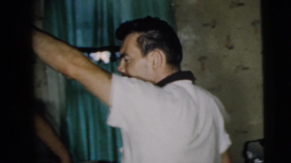 A man in a light shirt stands in the Camden room and raises his arm near green curtains and patterned walls.