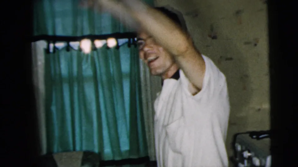 A smiling man in a white shirt waves toward the camera in a 1962 Camden private interior with green curtains behind him.