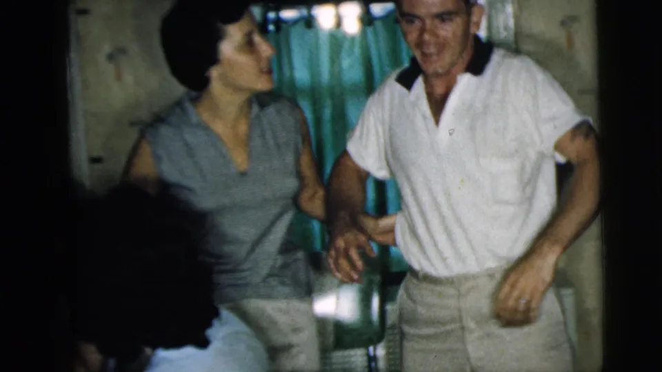 In a 1962 Camden home-movie frame, a man in a white polo stands beside a woman in a sleeveless top inside a private room.