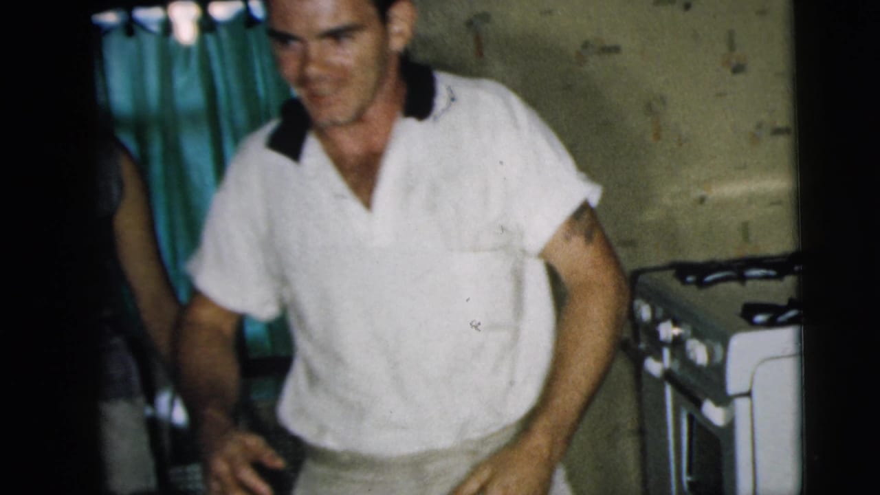 Man in a white polo shirt stands in a small kitchen near a white stove.