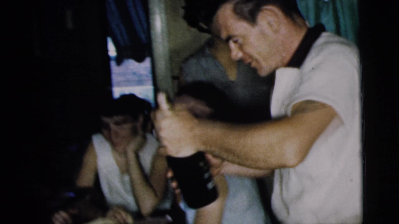 A smiling man holds a dark bottle while adults sit nearby in a dim 1962 Camden kitchen gathering.