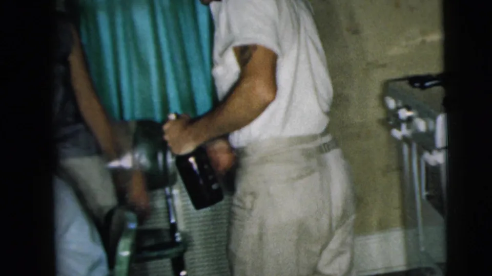 A man in light clothing turns with a dark bottle in a small Camden kitchen, with another adult partly visible beside him.