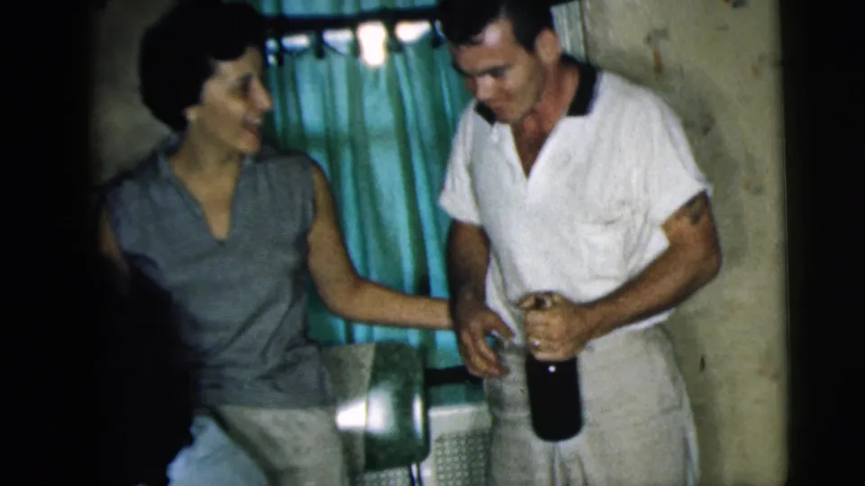 A woman smiles beside a man holding a bottle during a 1962 Camden kitchen celebration.