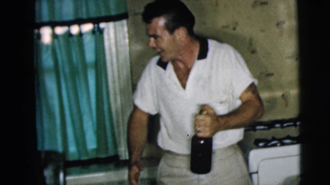 Man in a white shirt stands in a kitchen holding a dark bottle.