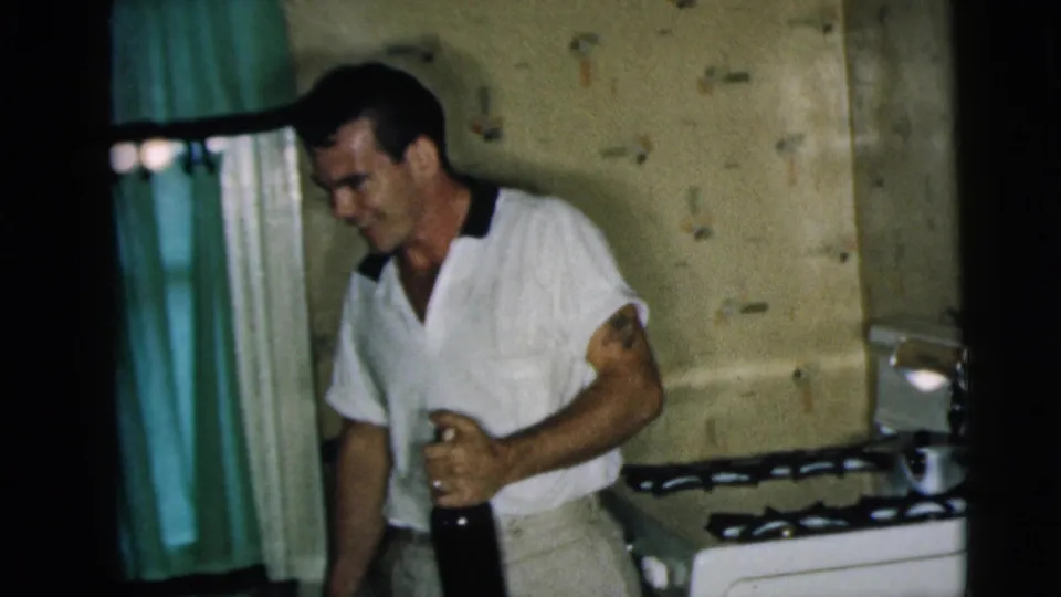 The man remains in the 1962 Camden kitchen, holding a dark bottle beside turquoise curtains, patterned walls and a white stove.