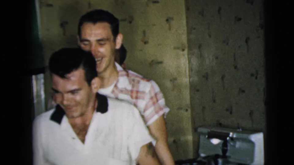 Two men smile in the Camden kitchen, with the foreground man holding a dark bottle and another man standing close behind him.