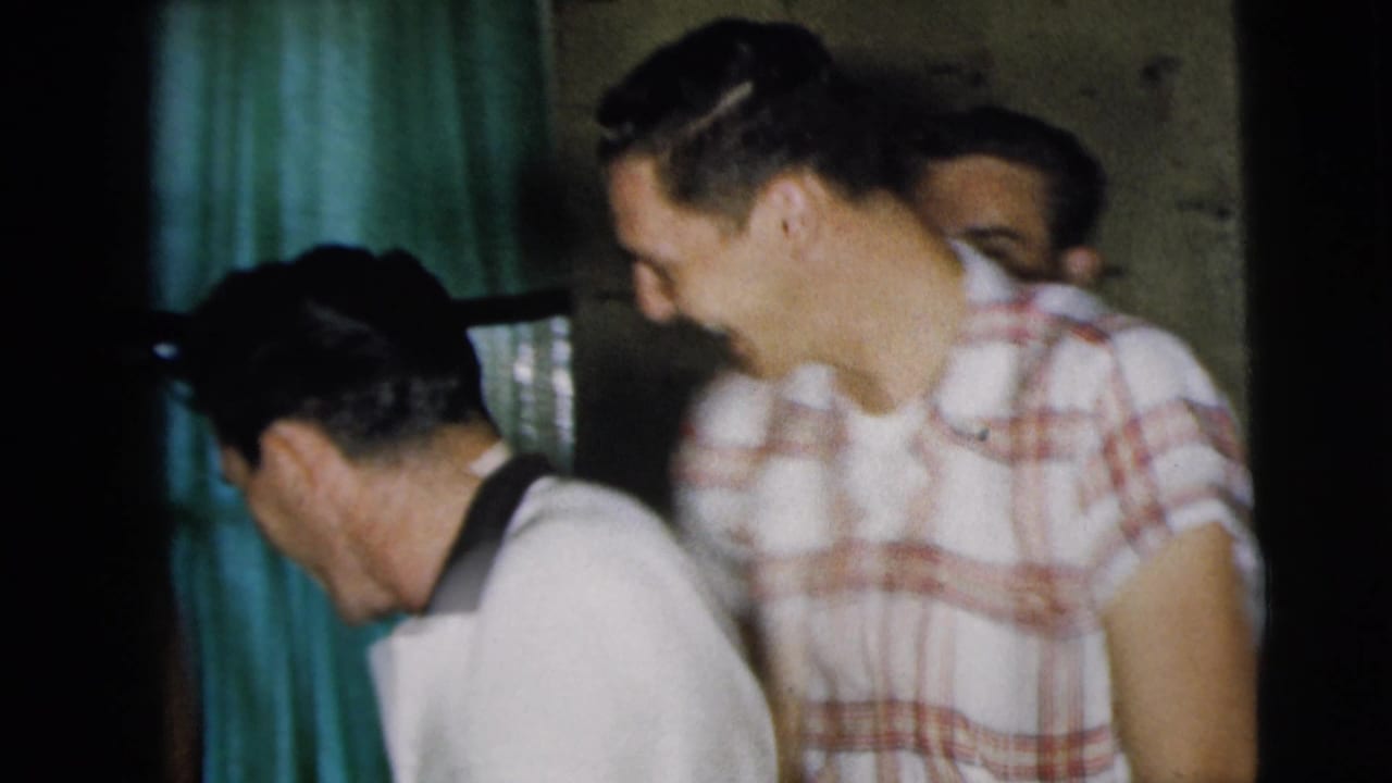 Young men in short-sleeved shirts stand in a dim home interior.