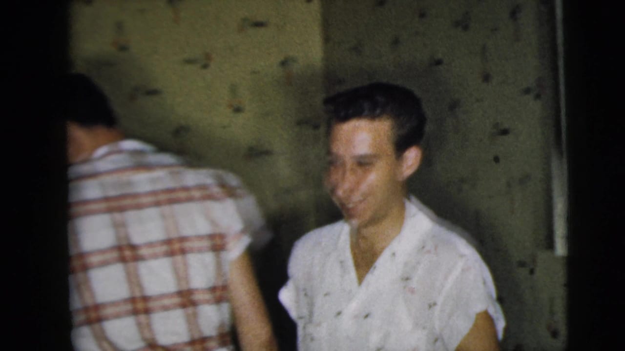 A smiling young Caucasian man stands in a patterned-wallpaper room as another man in a plaid shirt turns away.