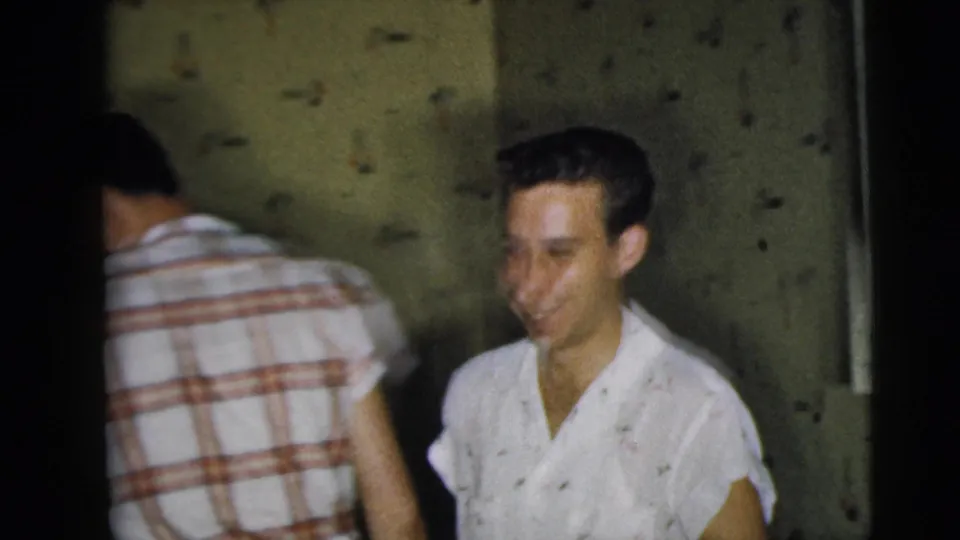 A smiling young Caucasian man stands in a patterned-wallpaper room as another man in a plaid shirt turns away.