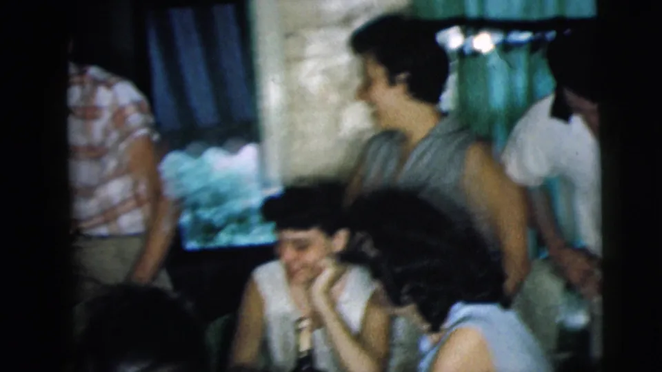 Young Caucasian adults sit and stand near a window in a Camden home, with one woman smiling at the table during the 1962 gathering.
