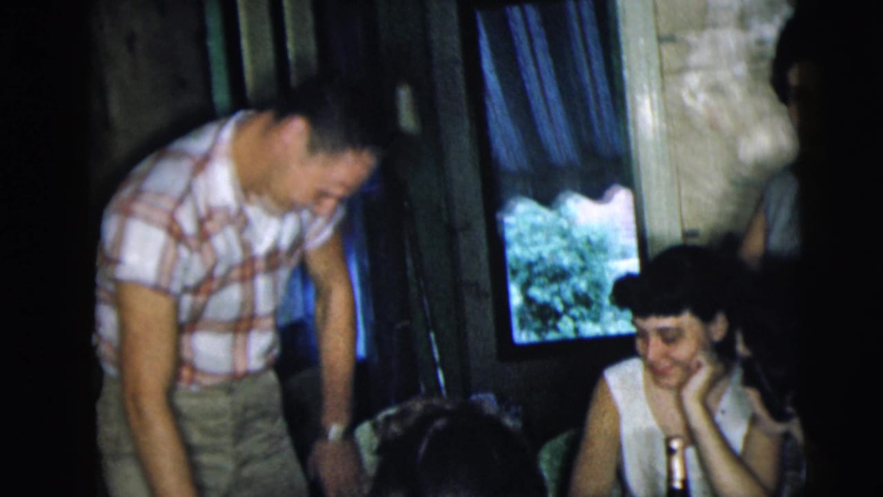 Young adults gathered in a dim room, with one man leaning near seated women at a table.