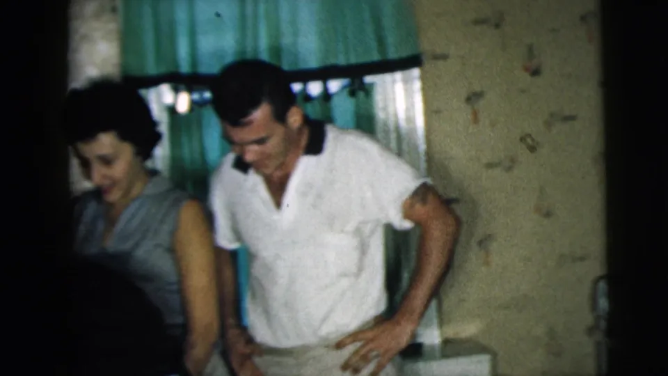 1962 Camden home-movie frame of a woman in a sleeveless dress and a man in a white shirt standing by seated friends.
