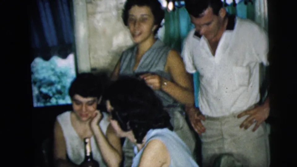 1962 Camden home-movie frame showing friends around a table, with a bottle visible and green curtains in the background.
