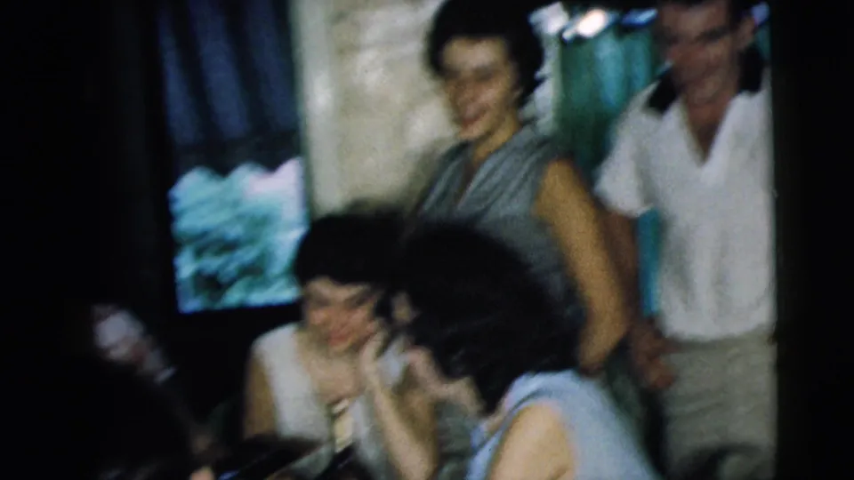 1962 Camden home-movie frame with soft motion blur as young adults continue socializing in a private room.