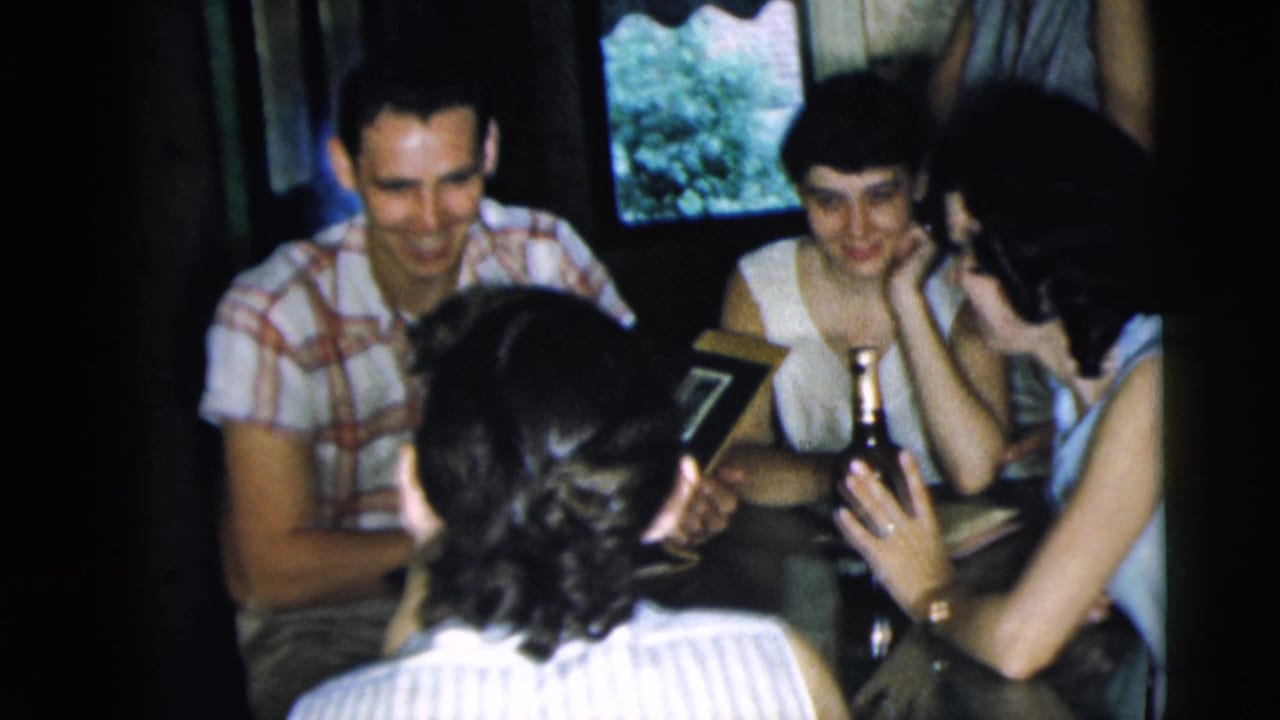In 1962 Camden home-movie footage, young adults sit close together around a table with a dark bottle and photo album.