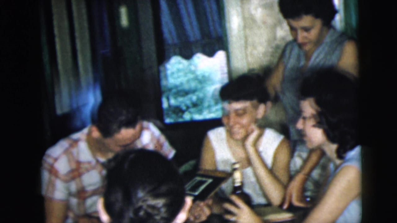 Young adults in a Camden, New Jersey interior smile and lean toward a table with bottles and a photo album in 1962 footage.