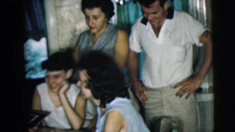 The home-movie frame shows adults gathered tightly in a private room, with one man standing beside seated women near the table.