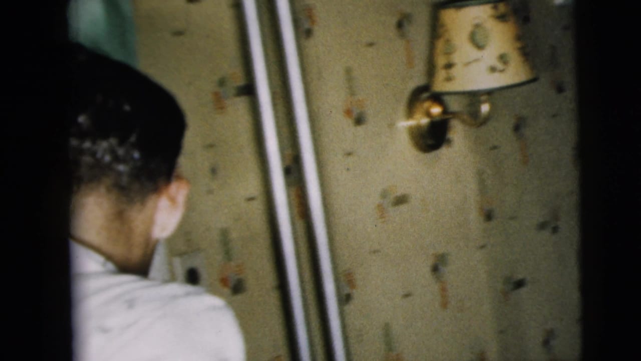 In a Camden, New Jersey home-movie frame from 1962, a man in a white shirt faces away near patterned wallpaper and a wall sconce.