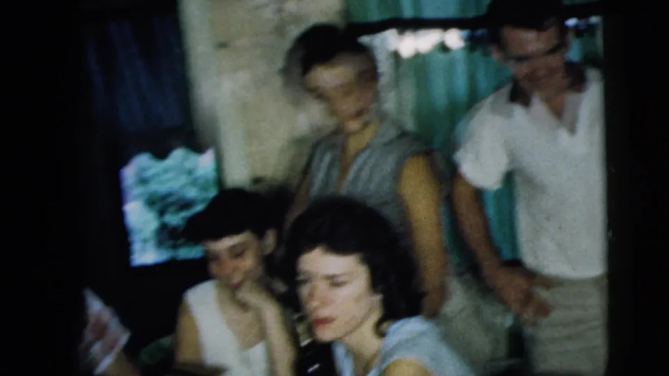 In a 1962 Camden interior, several adults gather close together near curtains, with seated women in the foreground and adults standing behind.