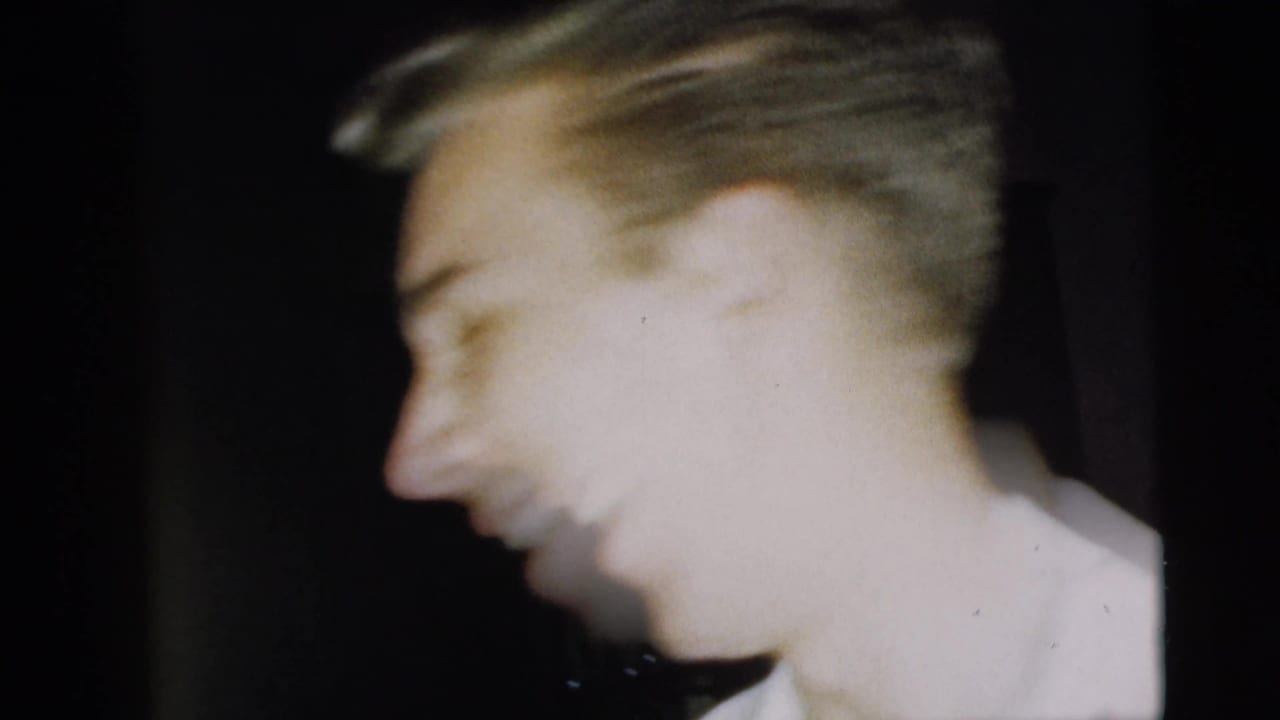 Blurred side view of a smiling man in a white shirt against a dark interior.