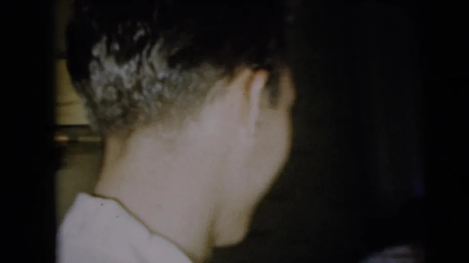 Rear view of a man in a white shirt, filmed close indoors during the Camden reunion sequence.