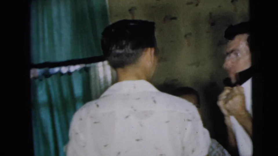 Group of men gathered indoors in Camden, New Jersey, with patterned wallpaper and a green curtain visible in the 1962 home-movie frame.