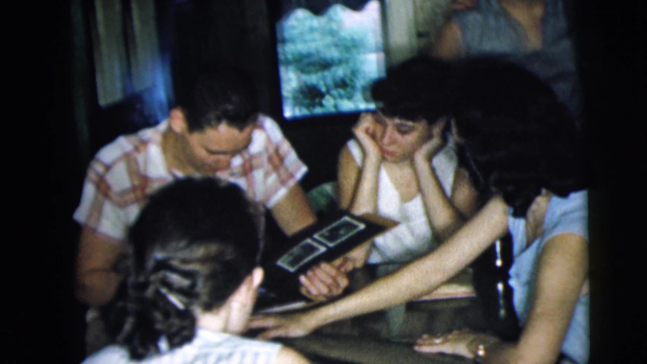 Young adults sit around a table looking at a photo album in a dim room.