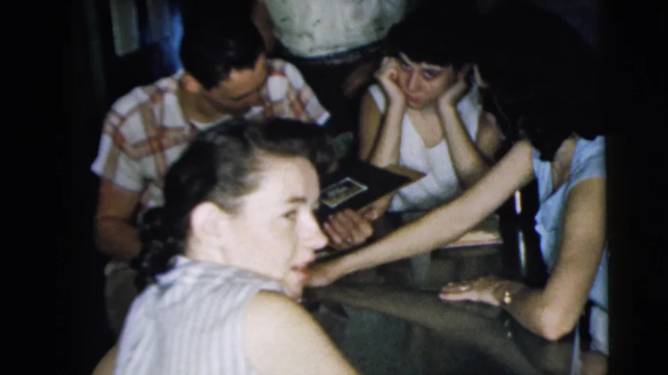 In the 1962 home-movie frame, one woman looks toward the camera as others around the table continue examining a photo album.