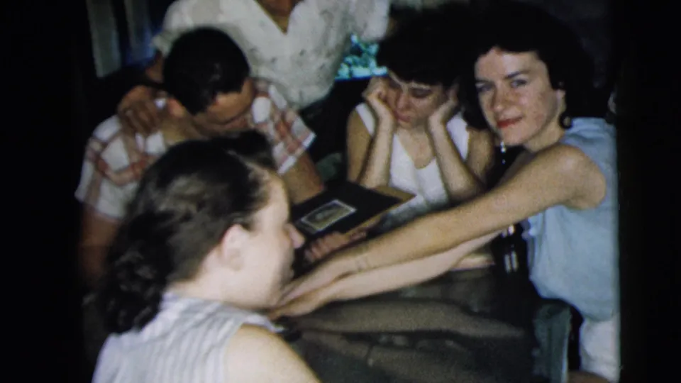 This Camden home-movie frame shows young adults gathered tightly around a table, with a photo album open as one woman looks toward the camera.