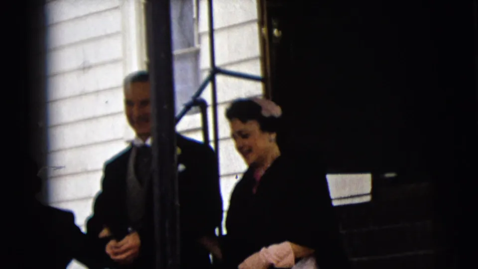 In Camden, New Jersey in 1962, an older couple in formal wedding attire appears near a white-clad building entrance.