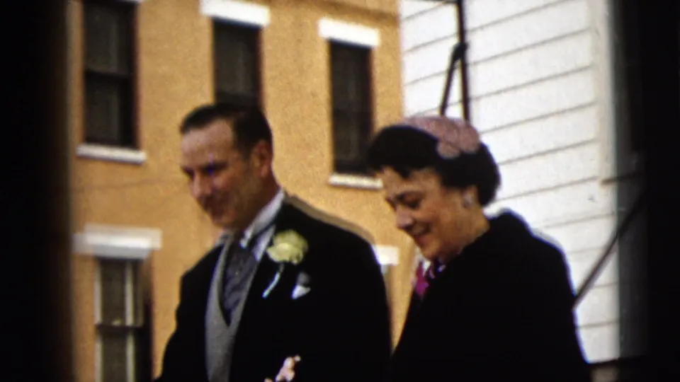 The older couple walks arm-in-arm outside the Camden building, with the groom's boutonniere and the bride's pink hat visible.