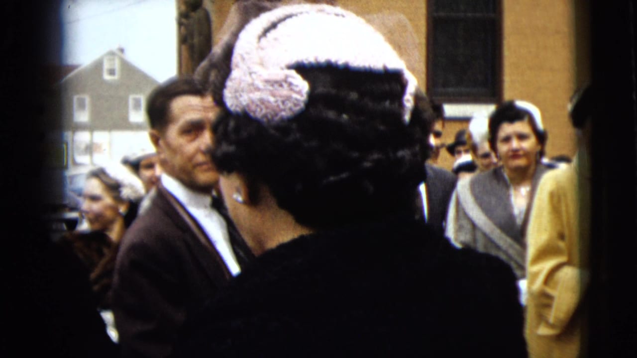 Wedding guests stand outside while a woman in a light hat faces away near a man in a dark suit.