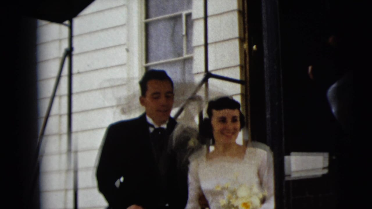 Bride and groom stand near exterior steps beside a white clapboard building.