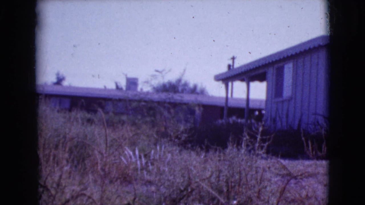 Small wooden house with a covered porch beyond dry grass and brush.