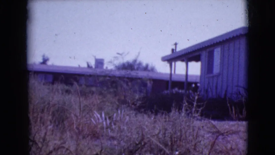 1964 8mm frame of a modest ranch-style house in Cottonwood, Arizona, seen past overgrown dry vegetation.