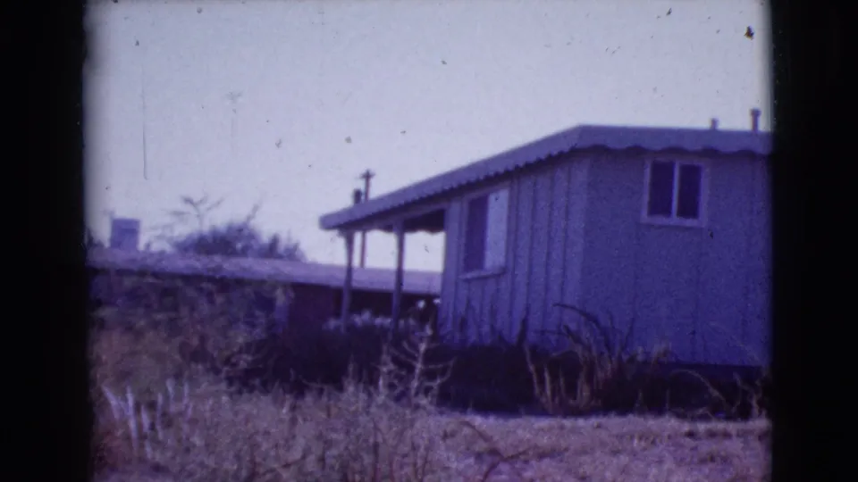 1964 home-movie view of a small Cottonwood house side, with a porch area, window, dry grass, and brush.