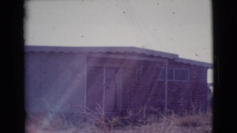 1964 Cottonwood, Arizona frame of a low residential exterior with a sliding door, brick section, overgrown dry grass, and soft color-shifted film exposure.