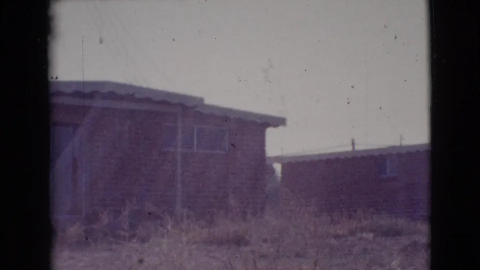 1964 Cottonwood, Arizona home-movie frame showing the side of a modest single-story house, sparse windows, dry yard, and adjacent building.