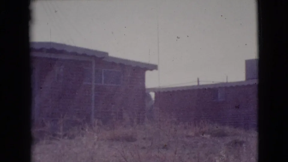 1964 Cottonwood, Arizona frame showing two modest low buildings in a row, dry foreground grass, simple windows, and soft 8mm texture.