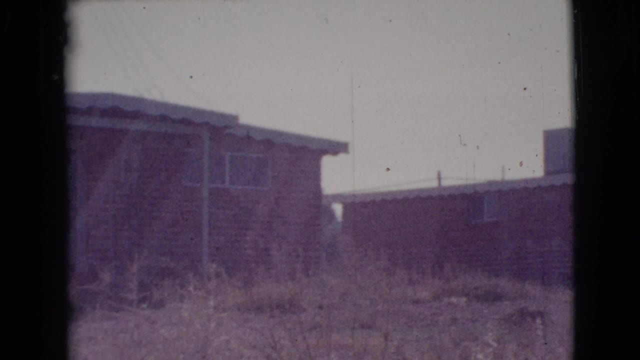 Modest residential houses seen across rough grass and dirt ground.