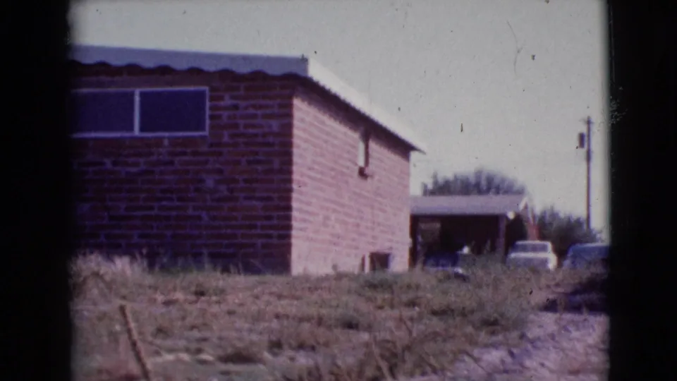 1964 Cottonwood, Arizona frame showing a brick residential exterior, small window, carport, parked cars, and scrubby ground.