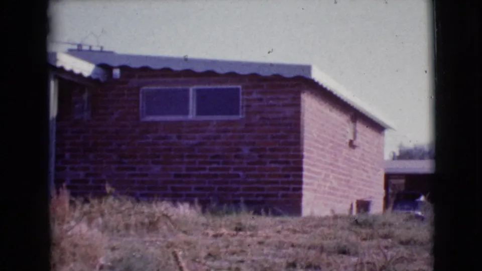 1964 Cottonwood, Arizona home-movie frame with the brick house shifted right, showing the roof edge, windows, dry grass, and adjacent shaded area.